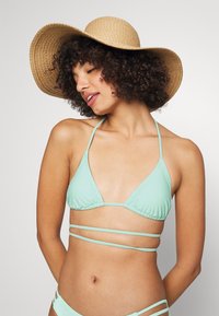 Piparmētru zaļa bikini ar trīsstūra formu un plānām siksnām, savienots ar plašu, salmu sauļošanās cepuri. Gluda tekstūra, minimālistisks dizains.