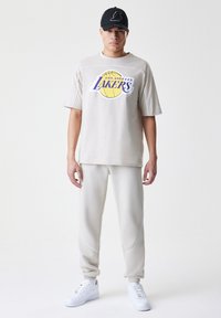 T-shirt oversized bege apresenta um gráfico dos "Los Angeles Lakers". Combinado com calças de moletão beige de corte tapered e ténis brancos. Inclui um boné preto.