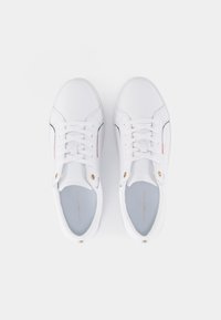 Un paio di sneaker bianche con lacci, con cuciture laterali sottili e occhielli dorati, viste dall'alto su uno sfondo bianco.