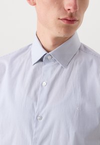 Lys blå og hvit stripete skjorte med klassisk krage, to knapper foran, og en liten logo brodert på brystet.