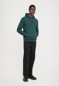 Felpa teal con cappuccio regolabile, tasca a marsupio frontale e logo. Abbinata a pantaloni neri e sneakers nere. Tessuto dalla consistenza liscia.