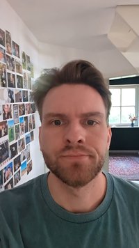 Homem com cabelo curto e escuro e barba, vestindo uma camisa azul-turquesa, está de pé em um corredor iluminado naturalmente, com uma colagem de fotografias na parede.