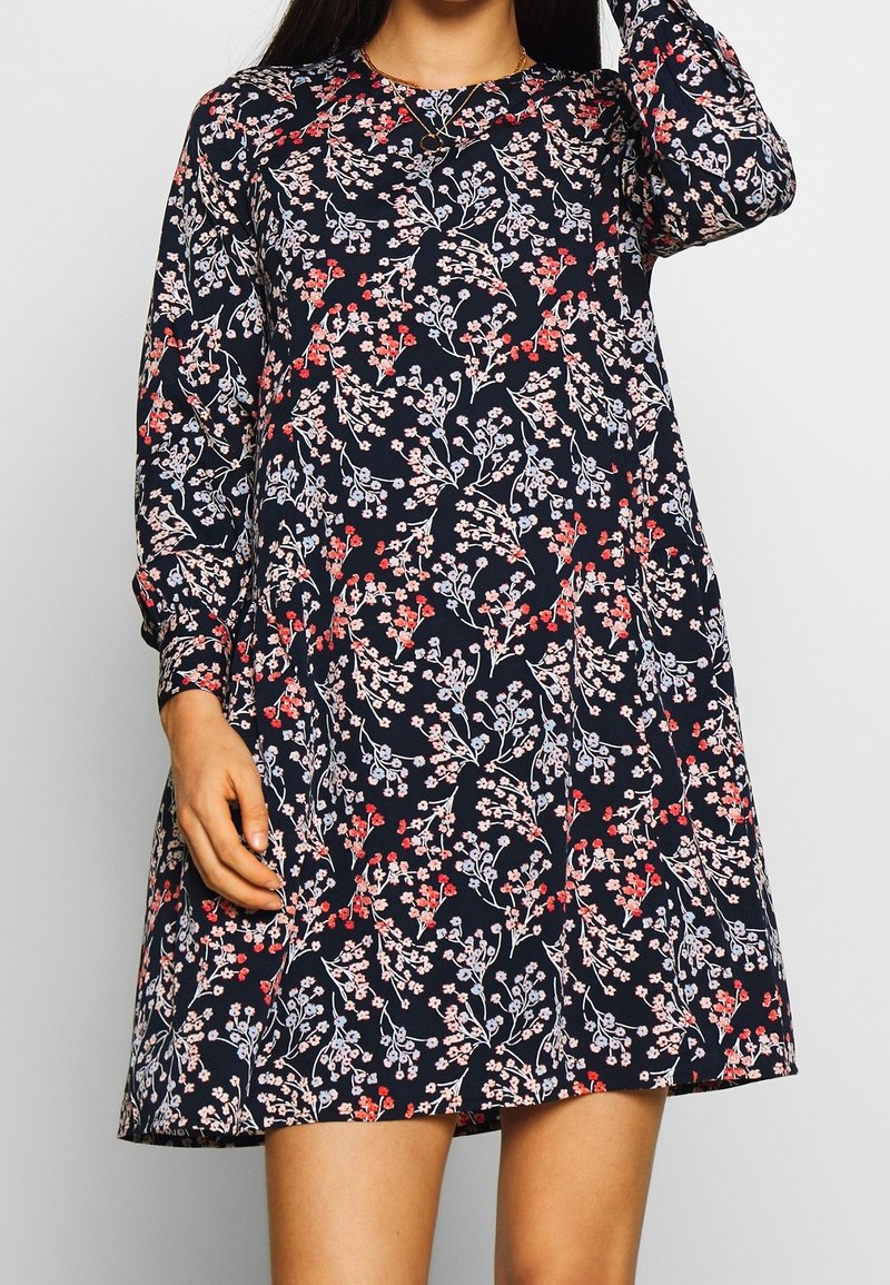 Femme portant une robe à manches longues bleu marine avec un motif floral rose et rouge, bras levé et main reposant près de la tête.