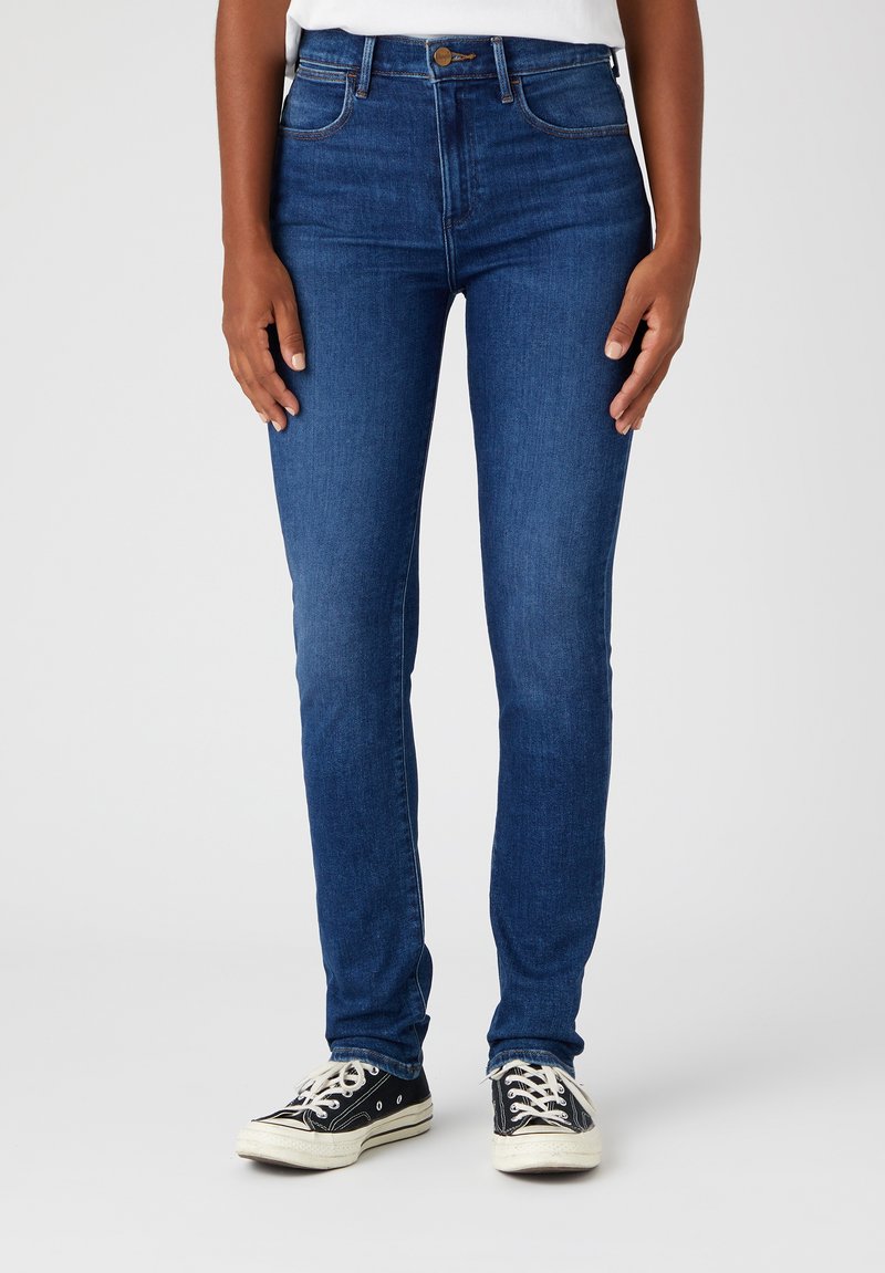 Jeans skinny blu scuro con vita alta, dotati di cinque tasche, passanti per cintura e dettagli di cucitura sottili. Indossati con sneakers nere a bassa altezza.