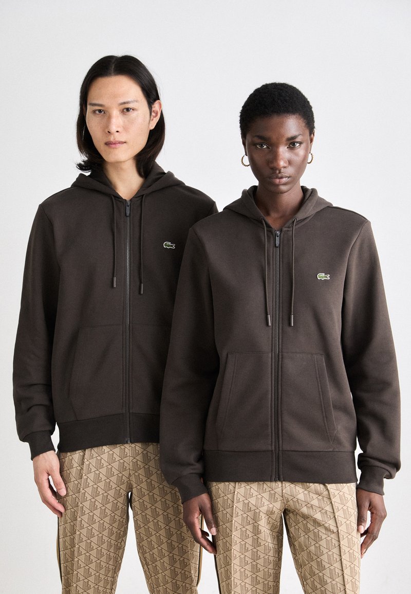 Hnedé mikiny na zips s kapucňou, prednými vreckami, sťahovacou šnúrkou a malým zeleným logom na hrudi, skombinované s vzorovanými béžovými nohavicami.