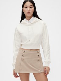 Bijela skraćena hoodie s vezicama, koja ima teksturirani logotip, u kombinaciji s bež mini suknjom s velikim gumbima sa strane.