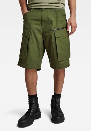 Person trägt olivgrüne knielange Cargoshorts mit mehreren Taschen und schwarze Schnürkampfstiefel mit schwarzen Socken.