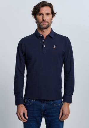 Hombre con cabello corto castaño y barba, vistiendo una camiseta polo de manga larga azul oscuro y vaqueros azules, de pie frente a un fondo liso.