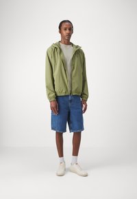 Giacca leggera verde chiaro con cappuccio, t-shirt grigia, pantaloni corti di jeans blu, sneakers bianche e calze grigie. Outfit casual con vestibilità rilassata.