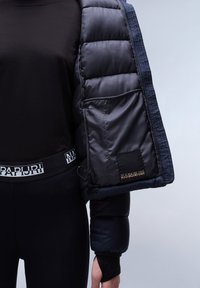 Puffjakke i mørk navy med quiltemønster, aftagelige ærmer og sort mærke. Bæret over en tætsiddende sort top og leggings.