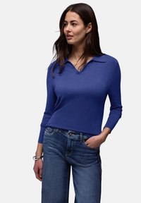 Femme aux longs cheveux bruns portant un pull bleu à manches longues avec un col en V et un jean bleu, une main dans la poche, regardant sur le côté.