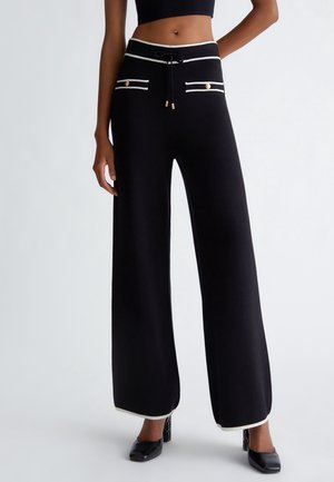 Mujer con pantalones anchos negros con ribete blanco, detalles de botones dorados, cintura con cordón y zapatos negros de tacón.
