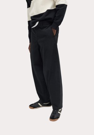 Osoba ubrana w czarne spodnie o szerokich nogawkach, czarno-białą bluzę oraz czarne trampki z beżowymi podeszwami na jednolitym tle.