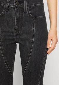 Čierne džíny z denimu s priliehavým strihom, s kontrastným šitím, prednými vreckami a stredne vysokým pásom. Ruka viditeľná na pravej strane.