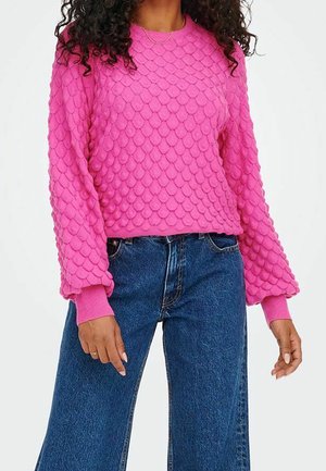 Kobieta ubrana w jasnoróżowy, teksturowany sweter z falistym wzorem oraz niebieskie, szerokie jeansy z wysokim stanem, stojąca na białym tle.