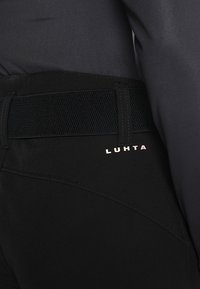 Pantalon de sport noir avec taille élastique et logo partiel "LUHTA" à l'arrière près de la taille, porté avec un haut noir.