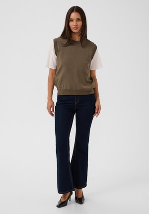 Femme aux longs cheveux foncés portant un pull sans manches vert olive par-dessus une chemise blanche, un jean bleu foncé et des chaussures noires à talons hauts.