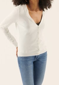 Vit stickad cardigan med V-ringning, med en rad knappar fram. Tillsammans med blå denimjeans som visar en figursydd stil.