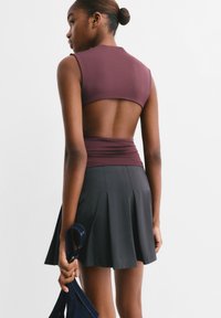 Lila aansluitend top met een open rug, gecombineerd met een geplooide zwarte rok. Het model houdt een marineblauwe tas vast, waarmee een gladde stofstructuur wordt getoond.