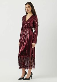 Abito bordeaux con paillettes, scollatura a V profonda, maniche lunghe e orlo con frange, abbinato a scarpe con tacco a punta nere.