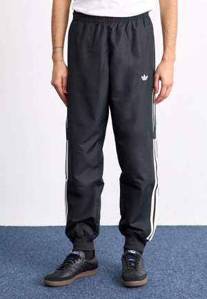 Persoon die zwarte Adidas trainingsbroek met witte strepen langs de zijkanten en zwarte sneakers met bruine zolen draagt, staand op een blauw tapijt.