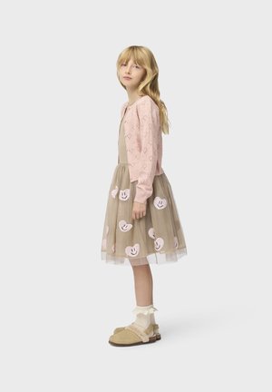 Ragazza giovane con lunghi capelli biondi che indossa un cardigan rosa lavorato a maglia e una gonna di tulle beige con motivi di cuori sorridenti rosa, in piedi di lato.