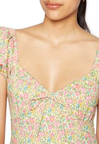 Vestido floral com fundo amarelo e padrões em rosa, azul e verde, decote em V e mangas bufantes com um detalhe de laço ao centro.