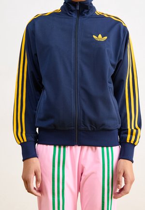 Person iført en mørkeblå lynlåstrøje med gule striber og logo, og lyserøde bukser med lodrette grønne striber, hænder afslappet ned langs siderne.