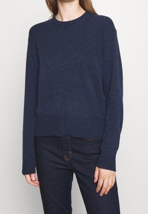 Femme portant un pull en maille bleu marine à manches longues et un jean bleu foncé, debout devant un fond clair uni.