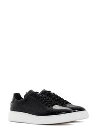 Zapatillas negras de caña baja con textura de cocodrilo, cordones negros y suelas de goma blanca gruesa, mostradas desde un ángulo frontal.