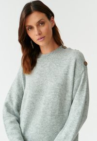 Pull gris clair en matière douce, avec un col rond et deux accents de boutons à l'épaule. Coupe décontractée avec des manches longues.