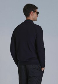 Marineblauer Strickpullover mit geripptem Kragen und Bündchen, ausgestattet mit einem Rundhalsausschnitt und einer glatten Textur. Das Modell trägt Sonnenbrillen und dunkle Hosen.