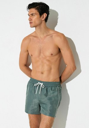 Muž bez košele s tmavými vlasmi, ktorý má na sebe zelené plavecké šortky s bielou šnúrkou, stojaci oproti jednoliatej bielej stene, pozerajúci sa doľava.