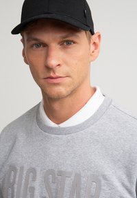 Ung mand med lys hud iført sort baseballkasket, grå sweatshirt med teksten "BIG STAR" og hvid skjorte med krave mod ensfarvet baggrund.