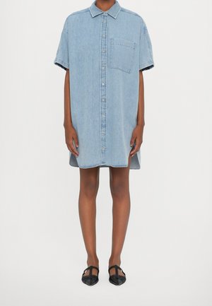 Osoba ubrana w jasnoniebieską, krótkorękawową sukienkę koszulową z denimu z guzikami z przodu oraz czarne, spiczaste płaskie buty.