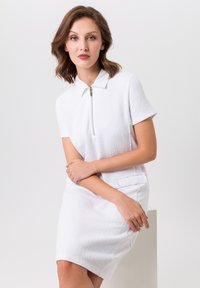 Witte jurk met korte mouwen van een textuurstof, polokraag en een rits aan de voorkant. Heeft een zijzak en een aansluitende silhouet.