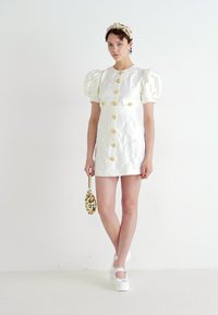 Λευκό φόρεμα με πούλιες και ραμμένο ύφασμα, χρυσά κουμπιά και λουλουδάτα σχέδια. Μοντέλο που κρατάει μια τσαντάκι με χάντρες και φοράει παπούτσια με πλατφόρμα.