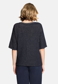 Femme aux cheveux blonds courts portant un haut noir à manches courtes en maille texturée ample et un pantalon foncé, vue de dos sur un fond blanc.