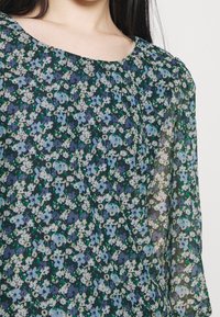 Květinová blůza s kulatým výstřihem, vyrobená z průsvitné látky v odstínech zelené, modré a bílé s malými květinovými vzory po celé ploše.
