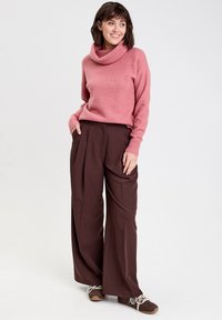 Pull en laine rose à col roulé avec des poignets en côtes, associé à un pantalon plissé ample de couleur marron foncé. Chaussures marron avec des lacets blancs.