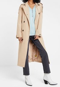Trench-coat beige avec de gros boutons, design croisé et épaulettes, porté sur un haut bleu clair, associé à un jean foncé et des bottines blanches.