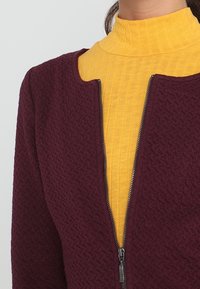 Burgunderfärgad texturerad jacka med dragkedja, lager på lager över en gul räfflad polotröja. Synlig sömnad och slätt tyg ger en intressant kontrast.