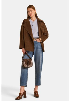 Frau trägt braunen oversized Mantel, hellblauen geknöpften Pullover, blaue Jeans, braune High Heels und hält eine kleine braune Handtasche.