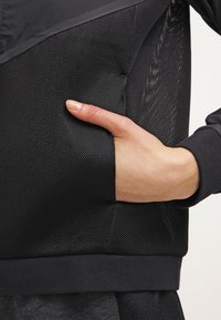 Zwarte sportieve jas met meshpanelen en een voorzak. De stof heeft een gestructureerde afwerking en een getailleerde taille met geribbelde manchetten.