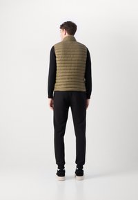 Colete acolchoado verde-oliva com gola alta, com um design acolchoado nas costas, usado sobre uma blusa preta de mangas longas e calças pretas.