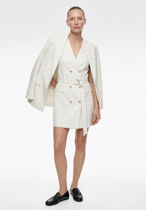 Kobieta ubrana w biały, prążkowany blezer zarzucony na ramiona oraz dopasowaną, dwurzędową sukienkę z paskiem, w czarnych mokasynach, stojąca na białym tle.