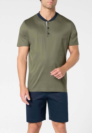Uomo che indossa una maglietta henley a maniche corte color verde oliva con collo e bottoni blu navy, abbinata a pantaloncini blu navy fino al ginocchio, in piedi davanti a uno sfondo bianco.