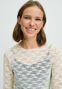 Krajková blúzka s dlhým rukávom v krémovej farbe, s kvetinovým vzorom a polo-priehľadným prekrytím. Mäkko na dotyk, s okrúhlym výstrihom a priliehavým strihom.