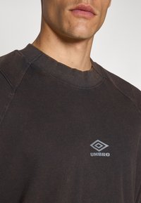 Tmavě hnědé bavlněné tričko s kulatým výstřihem a raglánovými rukávy. Na horní části hrudi je vytištěné světle šedé logo Umbro.