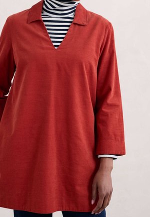 Personne portant une tunique longue à manches longues couleur orange brûlé par-dessus un col roulé rayé noir et blanc, debout devant un fond clair uni.